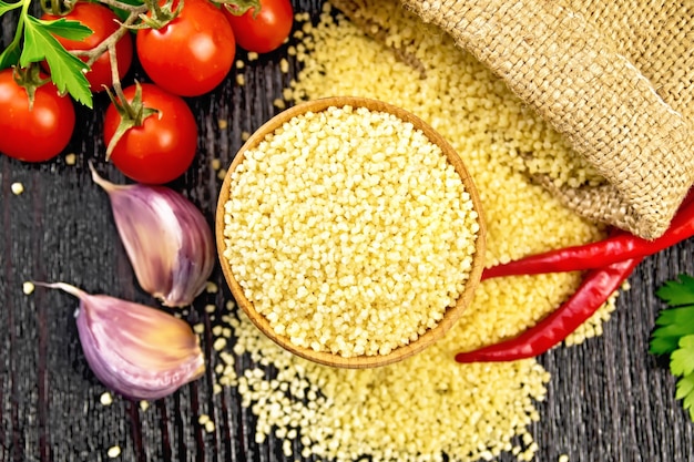 Raw couscous in a bowl and a sack of burlap, tomatoes, hot peppers, herbs and garlic on the background of dark wooden board from above