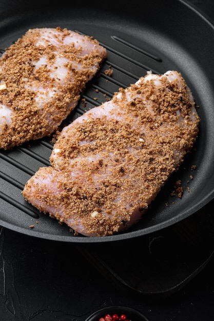 Raw crumbet chicken fillet with ingredient on cast iron skillet pan  over black background