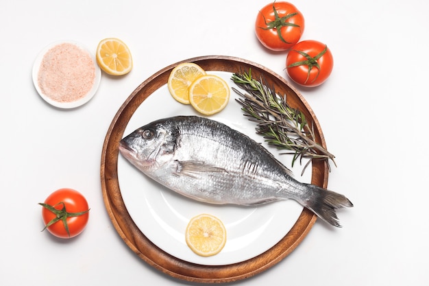 Сырая рыба дорада или позолоченный лещ подаются на белой тарелке на белой поверхности, плоская планировка, вид сверху