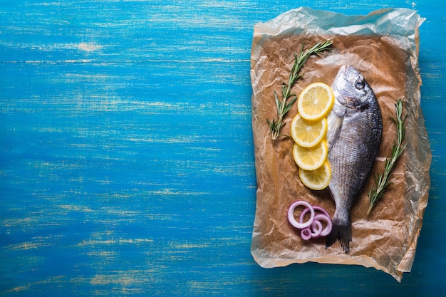 Raw Dorado fish prepared for cooking with slices of lemon, rosemary and onion on a blue background. Top view. Copy space.