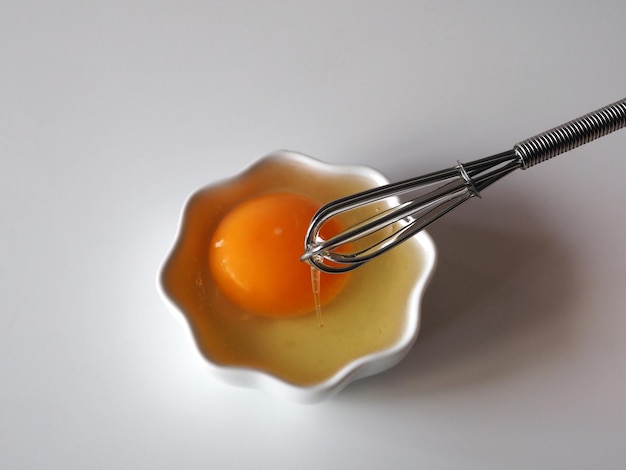 Сырое яйцо Яичный желток в белой миске и венчик для взбивания