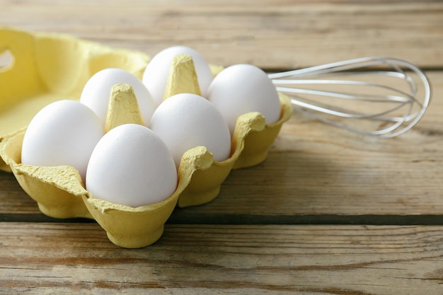 Raw eggs and whisk on wooden background