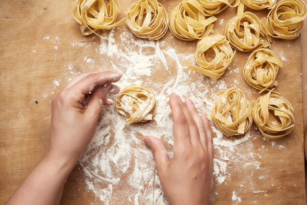 Foto pasta cruda di fettuccine e due mani femminili stanno cucinando la pasta su una tavola di legno marrone