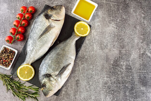 Raw fish dorado on black slate cutting board and grey concrete background with spices, tomato, rosemary, olive oil and lemon. Top view, flat lay with copy space for text