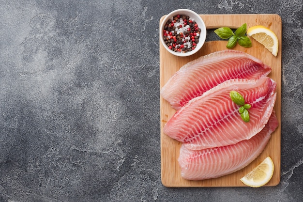 Raw fish fillet of tilapia on a cutting Board with lemon and spices. Dark table with copy space.