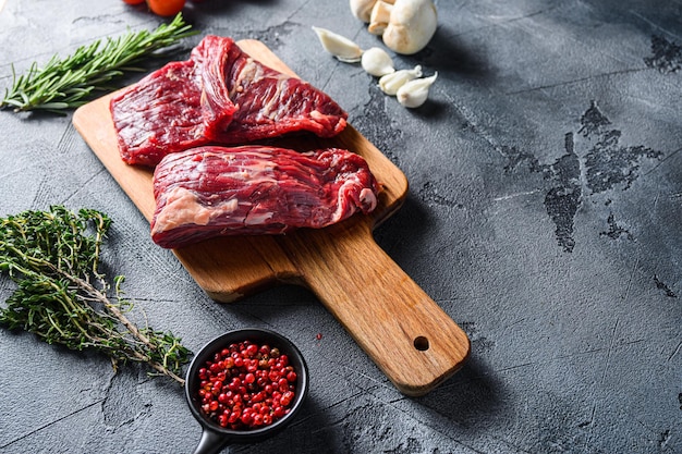 Raw flap steak flank cut with Machete, Skirt Steak, on woods chopping board, with herbs tomatoes peppercorns over grey stone surface background  side view space for text.