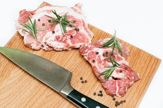 白い背景で隔離のまな板にローズマリー、コショウ、ナイフを添えた生の新鮮な牛肉ステーキは、家庭での料理をクローズアップします。