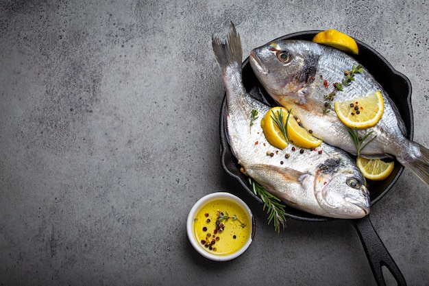 Raw fresh fish sea bream or dorado with lemon, thyme, rosemary and seasonings in cast iron skillet for cooking healthy meal or Mediterranean diet on rustic grey stone background above with copy space