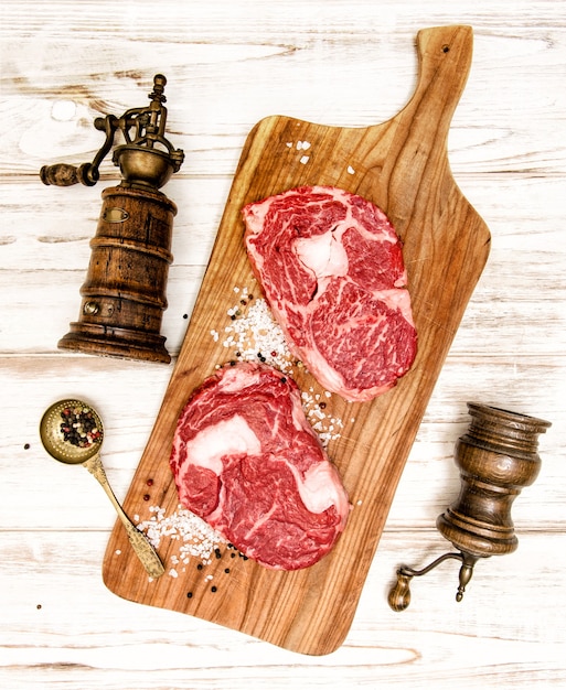 Стейк Рибай сырого свежего мяса с кухонной утварью на деревянном столе. Вилка и нож для приготовления пищи
