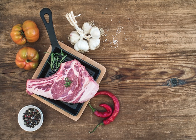 Raw fresh meat ribeye steak with pepper, salt, chili, garlic, spinach, heirloom tomatoes and rosemary in cooking pan over rustic wooden table