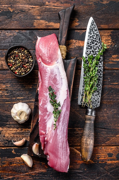 Raw fresh pork tenderloin meat on a cuuting board with cleaver. Dark wooden background. Top view.