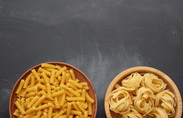 Raw fusilli and fettuccine pasta in wooden plates on a black table