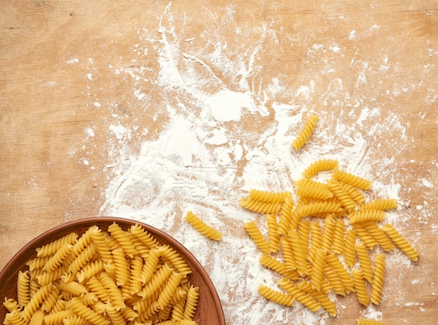 Foto pasta di fusilli cruda in un piatto marrone in ceramica su un tavolo di legno con farina bianca spruzzata vista superiore