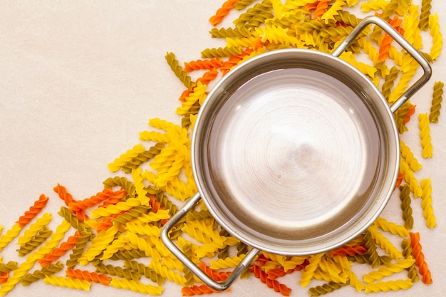 Raw fuzilli with water in pot. Multicolored pasta scattered over stone surface. Preparing for children, top view, close up.