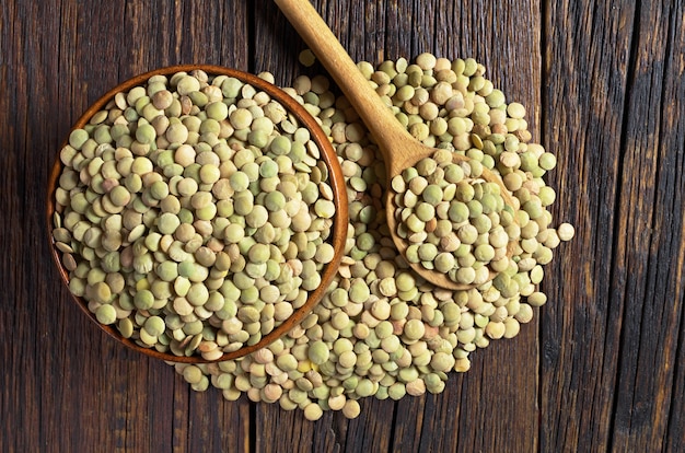 Raw green lentils in bowl and spoon near on dark wooden table, top view