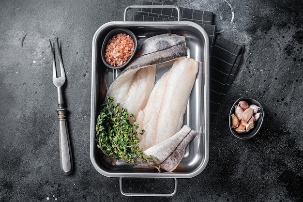 Raw haddock fish fillets, whitefish meat in kitchen tray with thyme. Black background. Top view.