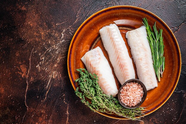 Raw Haddock loin fillet fish on rustic plate with thyme and salt. Dark background. Top view. Copy space.