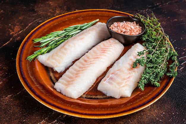 Raw Haddock loin fillet fish on rustic plate with thyme and salt. Dark background. Top view.