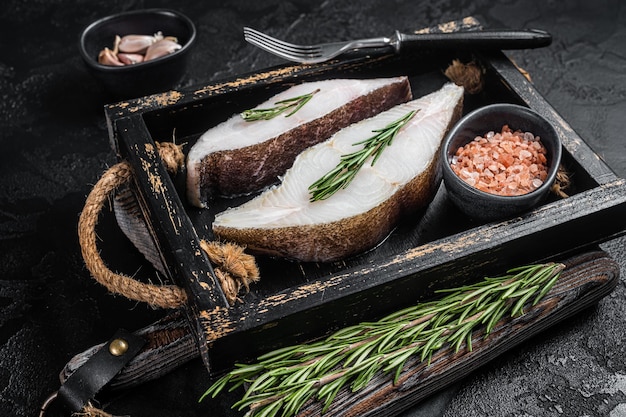 Raw halibut fish steak in wooden tray with herbs. Black background. Top view.