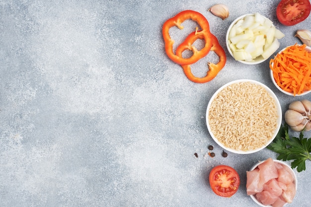 Raw ingredients for cooking pilaf, Brown rice and chicken fillet, grated carrots and chopped onions, pepper tomato garlic herbs and spices. Gray background, top view. copy space