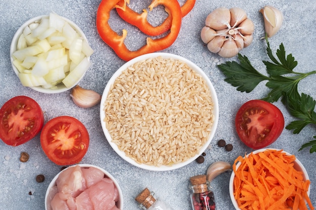 Raw ingredients for cooking pilaf, Brown rice and chicken fillet, grated carrots and chopped onions, pepper tomato garlic herbs and spices. Gray table, top view.