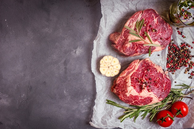 Raw juicy ribeye steaks with seasonings ready for roasting on baking paper. Rustic concrete background.