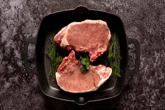 Raw juicy steaks with seasonings in a black pan ready for roasting on rustic concrete background. 