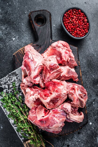 Raw lamb meat stew cuts with bone on wooden butcher board and\
cleaver. black background. top view.