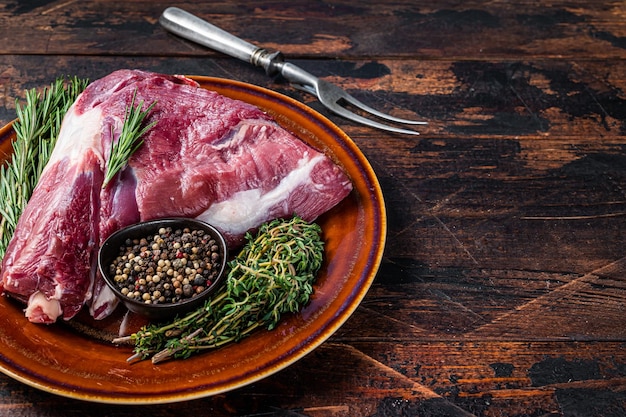 Raw lamb mutton thigh or leg with rosemary and thyme in rustic plate. Dark wooden background. Top view. Copy space.