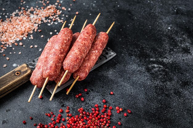 Raw lula or kofta kebabs skewers on a butcher cleaver with salt and pepper. Black background. Top view. Copy space.