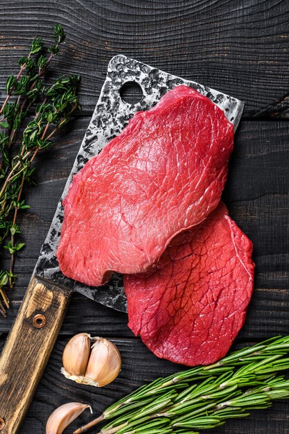 Raw marble beef meat fillet steak on butcher cleaver. Black wooden background. Top view.