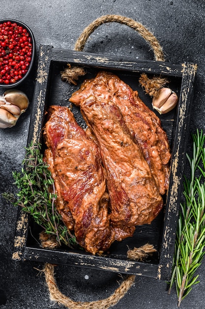 Raw marinated brisket steaks in a wooden tray with herbs.