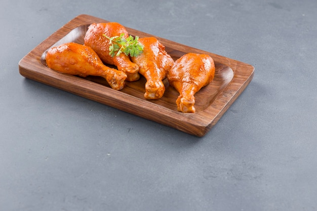 Raw marinated chicken legs or drum sticks arranged in a wooden\
base garnished with fresh coriander leaf which is placed on grey\
textured background.