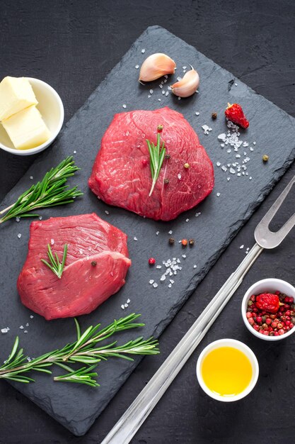 Raw meat beef steaks on black slate board, spices and ingredients for cooking. Top view