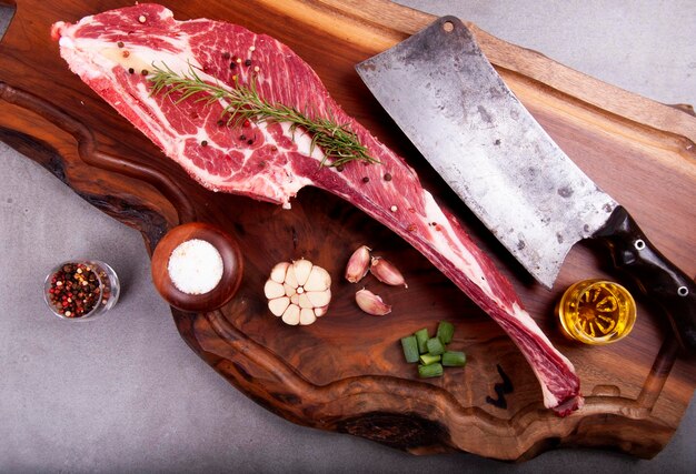 Raw meat cut short rib marbled with fat and bone on stone with spices and cleaver on wooden board top view