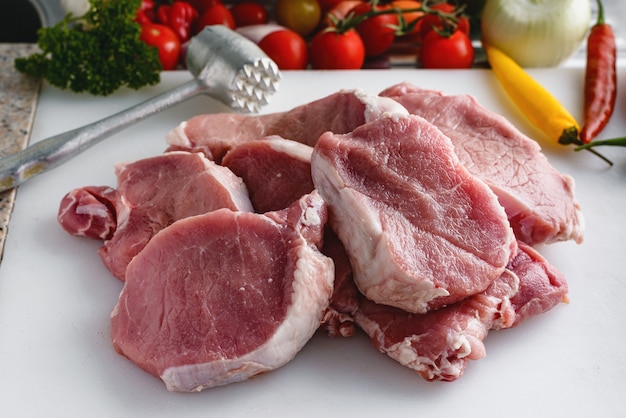 Raw meat, pork steaks on kitchen  table. Vegetables and meat hammer in the background.