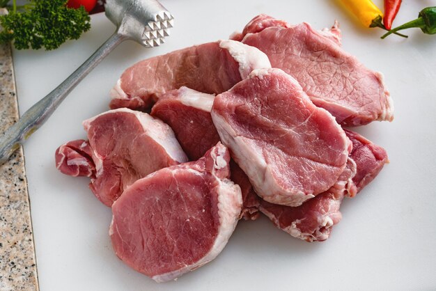 Carne cruda, bistecche di maiale sul tavolo della cucina. verdure e martello di carne in background.