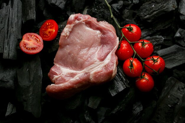 Raw meat steak on a background of charcoal raw steak with\
cherry tomatoes and rosemary on the coals