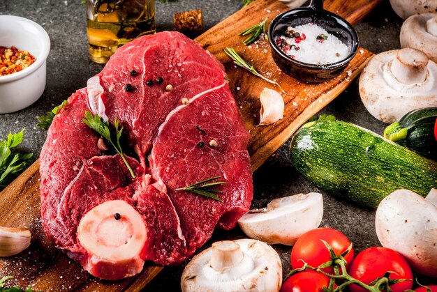 Raw meat with ingredients for dinner. Beef fillet, tenderloin, on cutting board, with salt, pepper, parsley, rosemary, oil, garlic, tomato, mushroom, zucchini.Black stone table