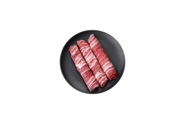 Raw minced meat wrapped in bacon with salt and spices or cevapcici on a dark concrete background