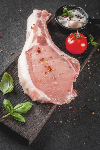 生の有機肉。食品調理の背景。骨付き豚肉。フライパン用のカツレツ。スパイスと食材バジル、ニンニク、木製のまな板copyspaceの黒いテーブルの上