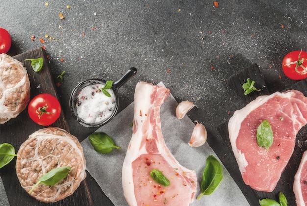 Raw organic meat. Selection of several types of red meat: pork cutlet on bone, pork steaks and chicken cutlets in grid of pork fat. With ingredients for cooking, gray stone table  