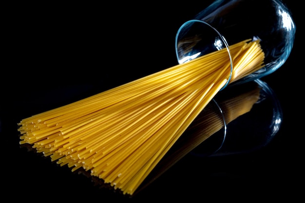 Photo raw pasta in glass jar wine glass in bucket raw pasta on black background front view raw pasta dropped from hand place for text yellow long spaghetti straws