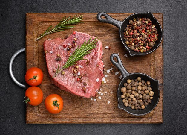 Raw piece of beef with spices pepper rosemary sprig salt and olive oil on a wooden board black background