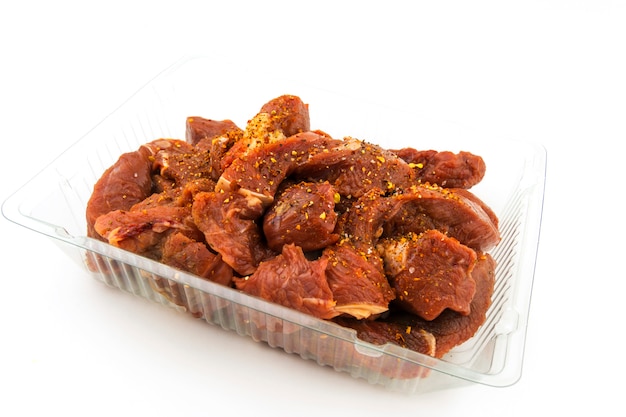 Raw pieces of beef, goulash in a plastic tray, sprinkled with spices. On white background.