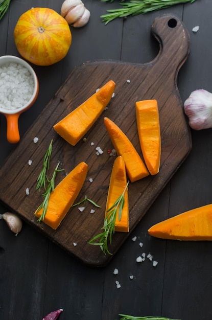 Photo raw pieces of pumpkin on a cutting board with rosemary, salt, spice on old dark table. vegetable prepared for baking in the oven. autumn home cooking