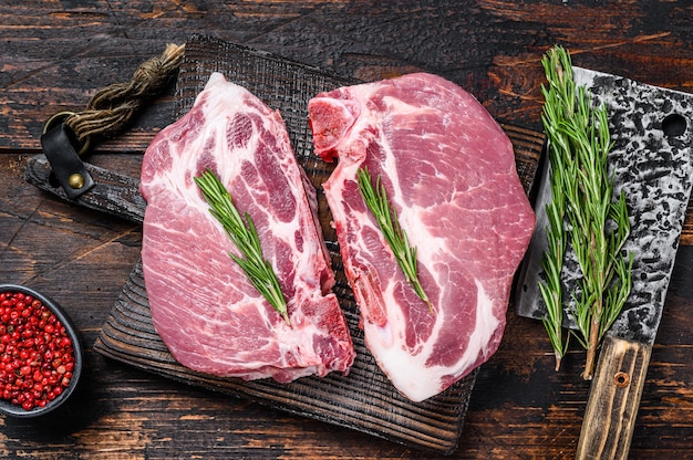 Raw pork loin steaks with herbs on a cutting board with meat cleaver. Dark wooden background. Top view.