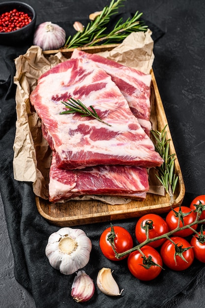 Raw pork meat ribs with ingredients for cooking rosemary and garlic in a wooden bowl. Black background. Top view