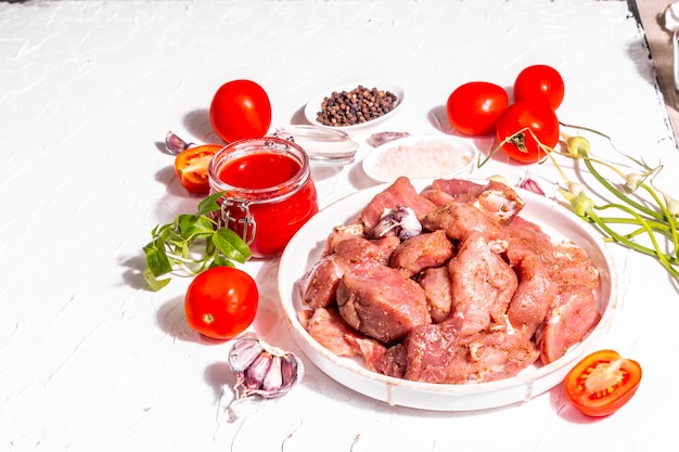 Raw pork pieces, tomatoes, spices, and herbs. Ingredients for cooking healthy food on a white putty background. Culinary process concept, trendy hard light, dark shadow, copy space