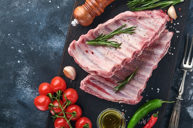 Raw pork ribs or fresh uncooked meat with spices on black wooden tray with paprika, garlic cloves and herbs. Dark textured background with copy space for text.